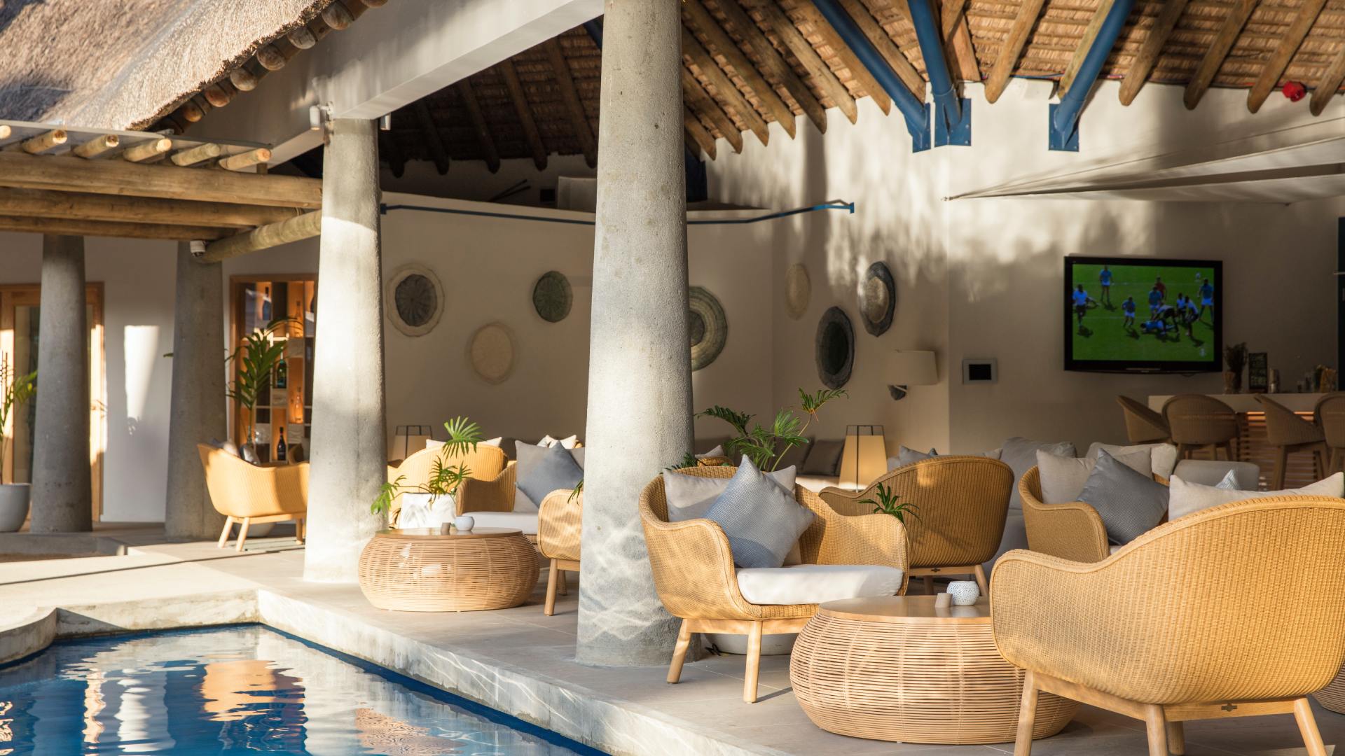 A serene pool scene at C-Beach Club Mauritius, complete with wicker chairs and a television for leisure activities.