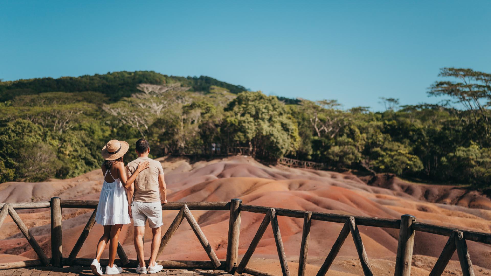 Two people on a wooden bridge, looking at the striking, colorful mountains of the Seven Colored Earth in Mauritius.